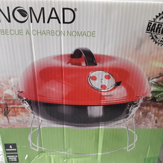 A red and black BE NOMAD charcoal barbecue grill in its original retail packaging, featuring a green base on the box with product imagery of food and herbs.