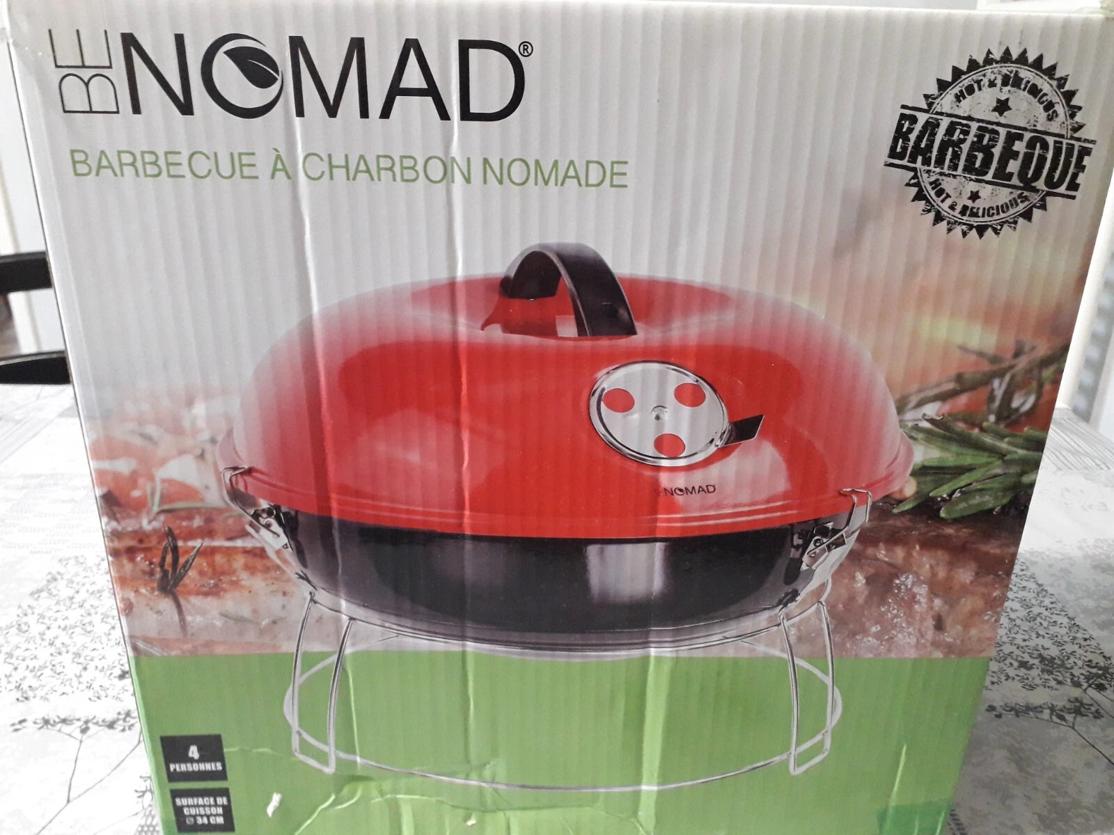 A red and black BE NOMAD charcoal barbecue grill in its original retail packaging, featuring a green base on the box with product imagery of food and herbs. -  Be Nomad Transportable Charcoal Barbecue Grill -  Abouche3le